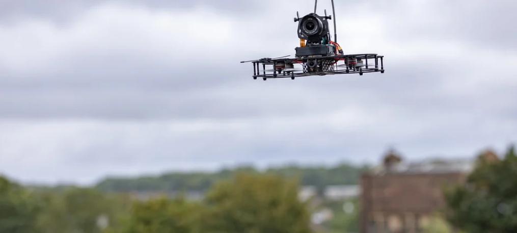 Eine Drohne fliegt in der Luft. Im Hintergrund sind Bäume und ein Haus zu sehen.