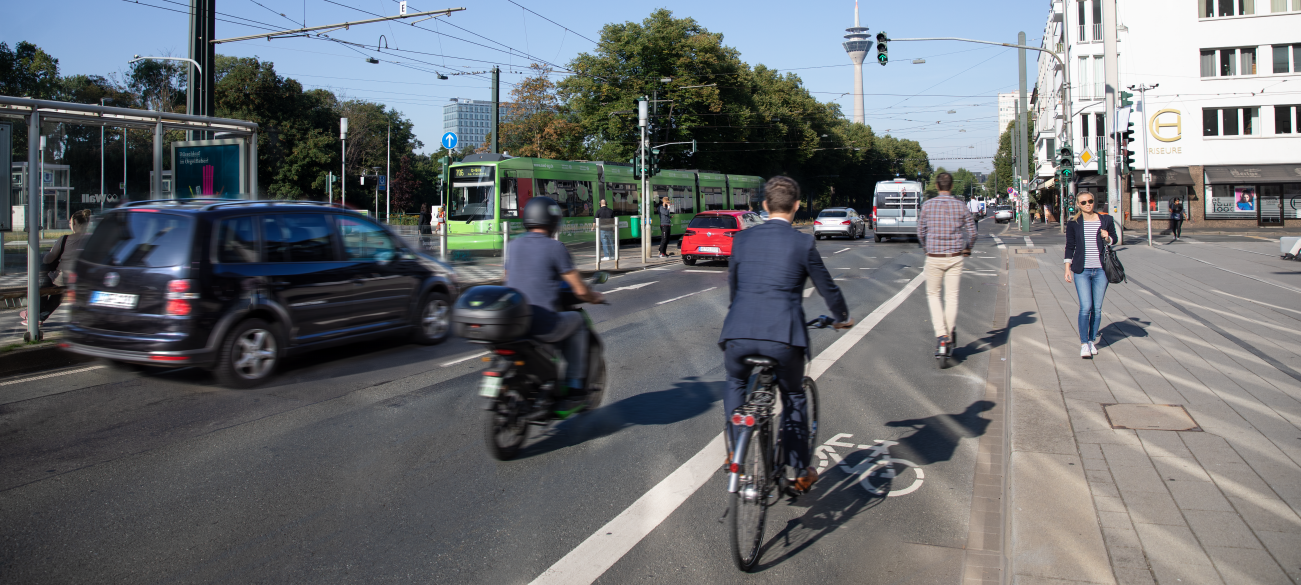 Das Foto zeigt verschiedene Formen der Mobilität am Graf-Adolf-Platz. Neben der Bahn fahren Autos. Außerdem sind Radfahrer auf einem Fahrradstreifen unterwegs und Fußgänger laufen über den Bürgersteig.