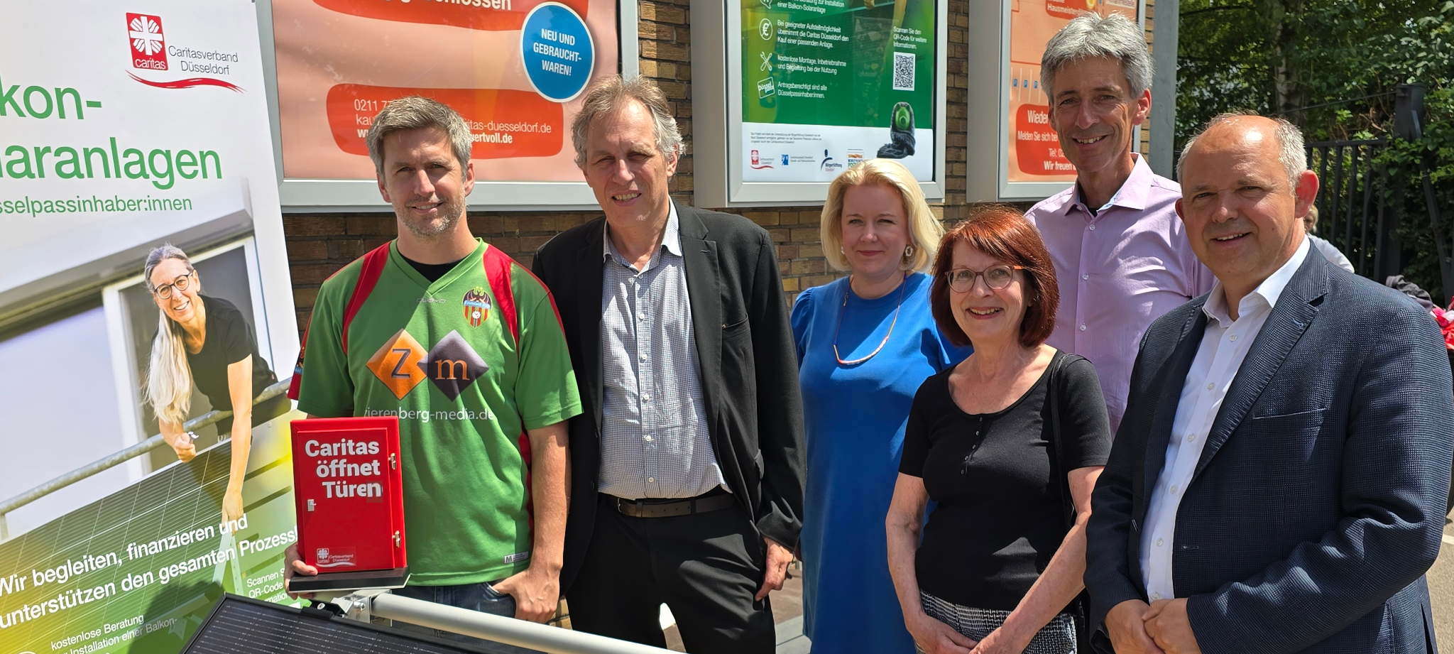 Sechs Menschen der Stadt, Caritas, Bürgerstiftung und Deutschen Postcode Lotterie stehen hinter einem Solarpanel. Einer von ihnen hält einen Flyer hoch.