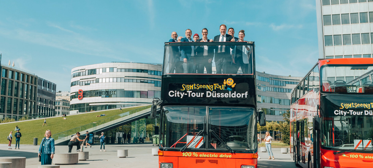 Blick auf die neuen elektrischen Hop-on-hop-off-Busse. Im offenen Oberdeck stehen der einige Personen und schauchen nach unten in die Kamera. Im Hintergrund ist der Kö-Bogen zwei zu sehen.