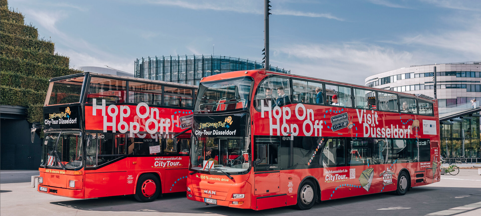 Blick auf zwei elektrische Hop-on-hop-off-Busse. Die Busse sind rote Doppeldecker-Busse mit Düsseldorf-Symbolen.