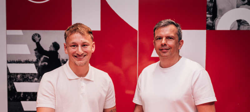 Sascha Risch und Sportvorstand Samir Arabi stehen vor einer roten Wand. Auf einer roten Theke liegt Risch' Vertrag. Er hält einen Stift in der Hand, beide lächeln.