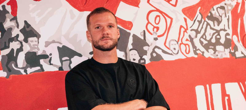 Der neue Fortuna-Mittelstürmer Eric Hottmann steht mit verschränkten Armen vor einer Wand, die mit rot-weißen Fortuna-Motiven geschmückt ist.