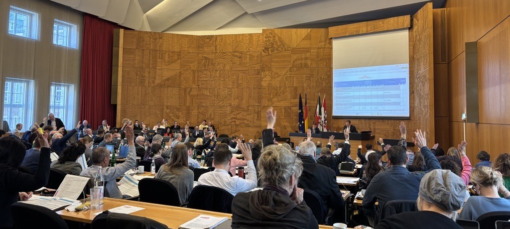 Der Rat der Stadt stimmt über den Haushalt 2026 ab: Viele Menschen sitzen in einem Saal und schauen nach vorne, einige haben die Hand gehoben.