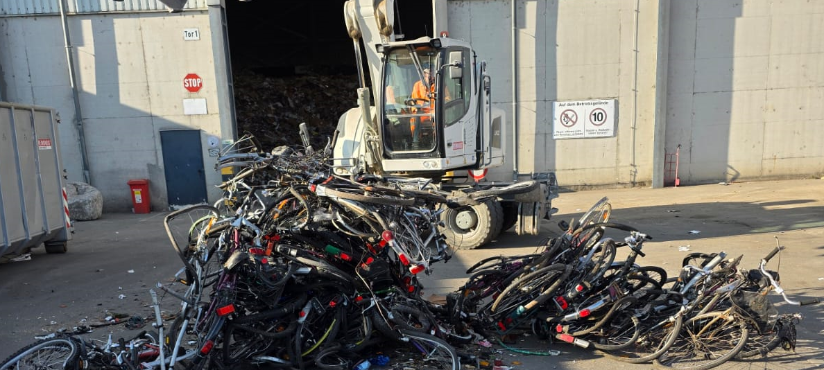 Auf einem Schrottplatz liegen viele kaputte Fahrräder auf einem Haufen. Im Hintergrund befindet sich ein Bagger.
